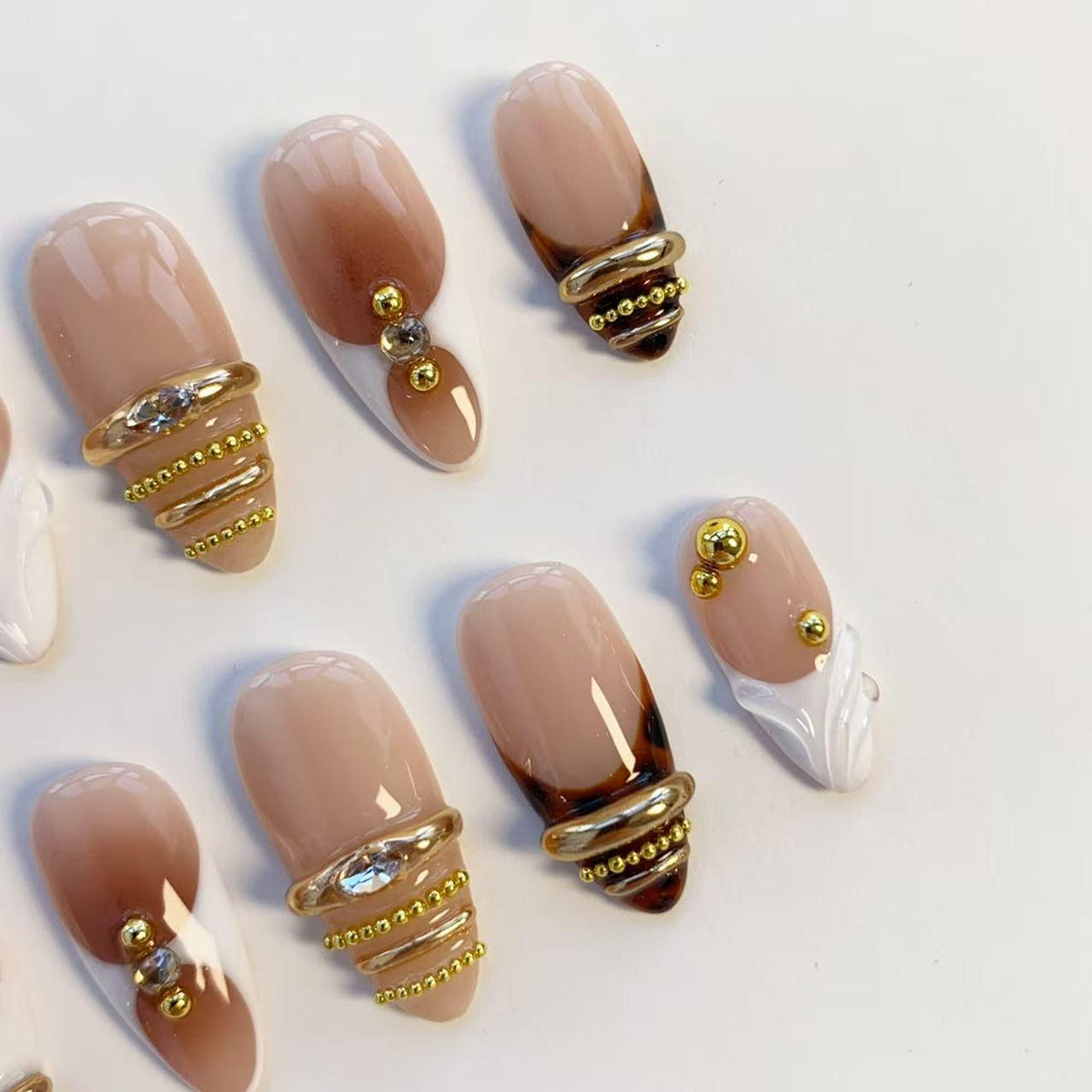 Oval-shaped artificial nails in various designs. Some nails feature a nude base with brown tips, while others have gold ring accents with rhinestones or small gold beads. A few nails have white tips.