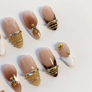 Oval-shaped artificial nails in various designs. Some nails feature a nude base with brown tips, while others have gold ring accents with rhinestones or small gold beads. A few nails have white tips.