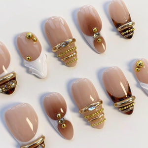A collection of artificial nails with a variety of designs. Some nails feature a nude base with gold embellishments, including rings and small beads. Others have a brown ombre effect or white tips, with additional gold accents.
