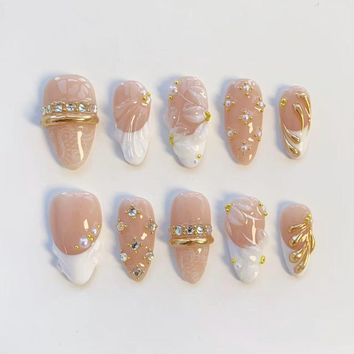A set of ten artificial nails in various designs. The nails feature a light pink base with white tips, gold accents, and rhinestones. Some nails have floral patterns, while others have gold bands with rhinestones.