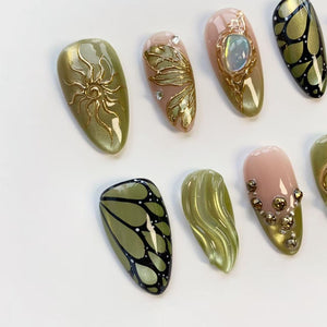 A collection of almond-shaped, painted fingernails. Designs include gold sunbursts, butterfly wings, and floral patterns. Colors include olive green, gold, black, and pink. Some nails feature rhinestones and iridescent accents.