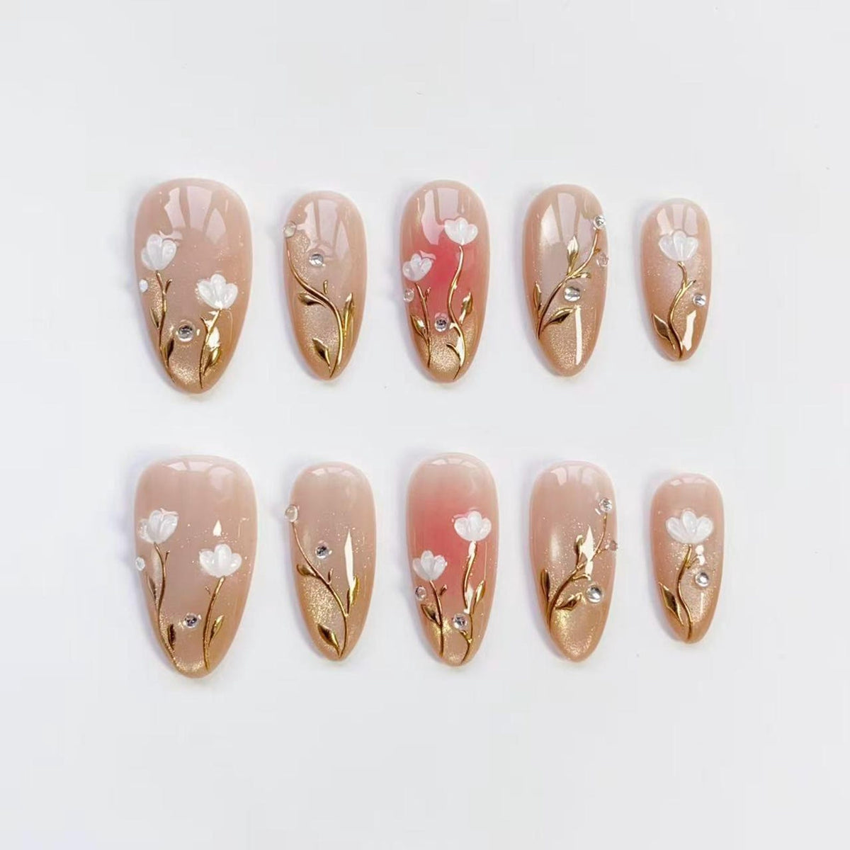 Set of ten oval-shaped false nails. Each nail features a nude base with a gold gradient, adorned with white floral designs, gold vines, and small rhinestones. The nails are arranged on a white surface.