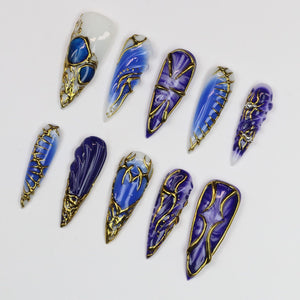 A collection of ten long, pointed false nails. Each nail features a design with shades of blue and gold. Some nails have a marbled effect, while others have intricate gold wire detailing.