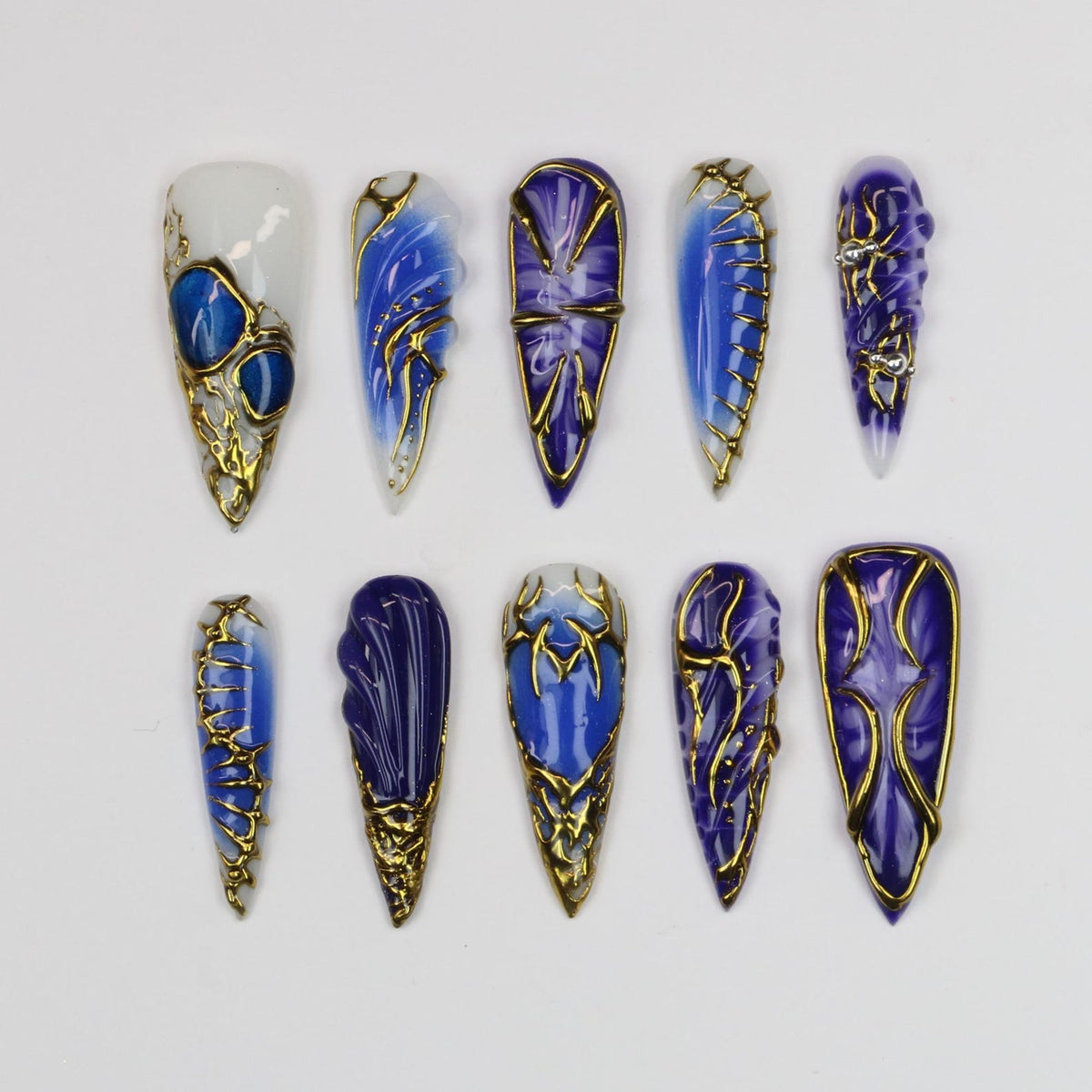 A collection of ten long, pointed artificial nails. Each nail features a unique design with shades of blue and gold accents. Some nails have a white base, while others are a solid blue. The gold accents create an ornate, elegant look.