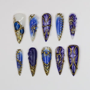 A collection of ten long, pointed artificial nails. Each nail features a unique design with shades of blue and gold accents. Some nails have a white base, while others are a solid blue. The gold accents create an ornate, elegant look.