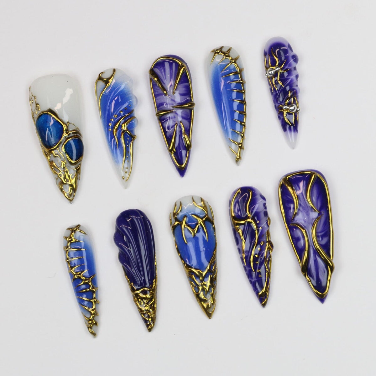 A collection of ten long, pointed artificial nails. Each nail features a unique design with shades of blue and gold accents. Some nails have a marbled effect, while others have intricate gold filigree.