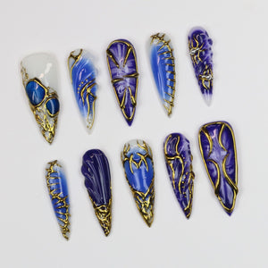 A collection of ten long, pointed artificial nails. Each nail features a unique design with shades of blue and gold accents. Some nails have a marbled effect, while others have intricate gold filigree.