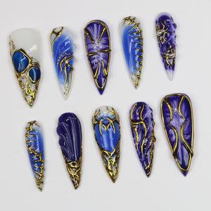 A collection of ten long, almond-shaped artificial nails. Each nail features a unique design with shades of blue and gold. Some nails have intricate gold filigree, while others showcase marbled or swirled blue patterns.