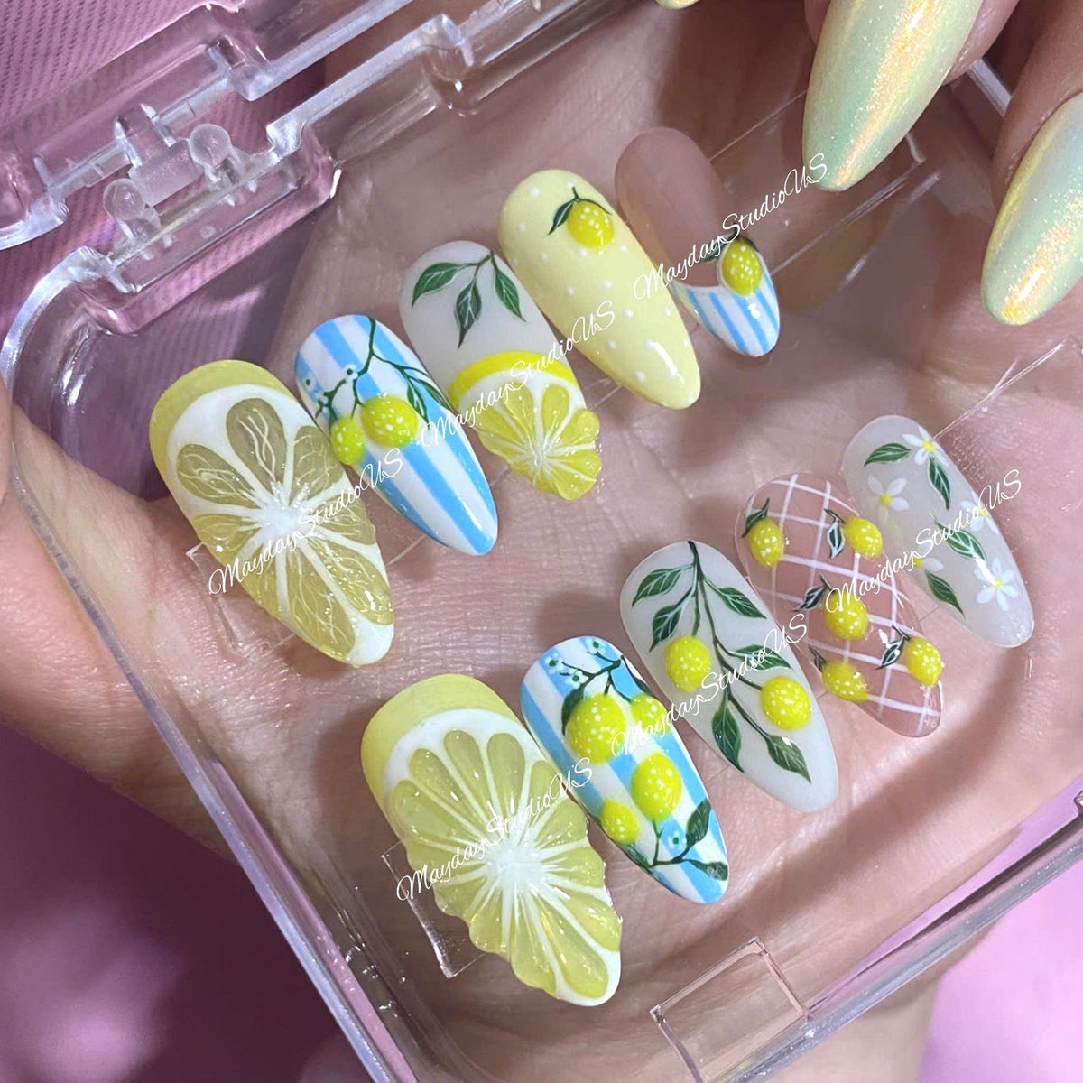 A set of artificial nails with various designs, including lemon slices, lemons with leaves, and blue stripes. The nails are in a clear plastic case. The text "Mayday Studio US" is visible on some nails.