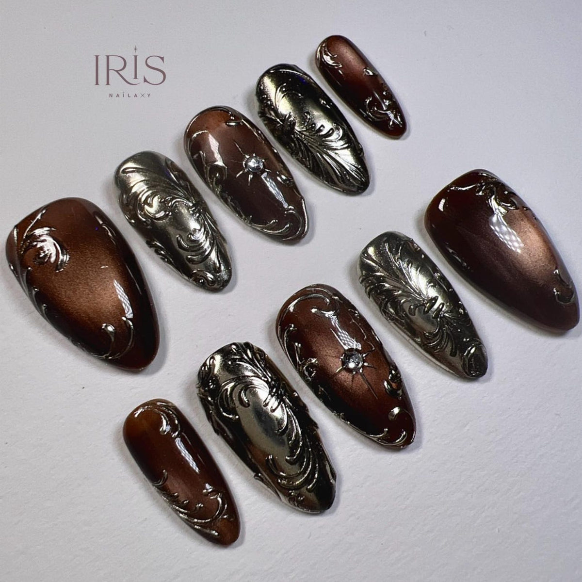 A set of press-on nails in various sizes, featuring a metallic silver and brown color scheme. The nails have ornate, embossed designs, including swirls and floral patterns. Some nails have a small, clear gem. The brand name "IRIS NAILART" is visible.
