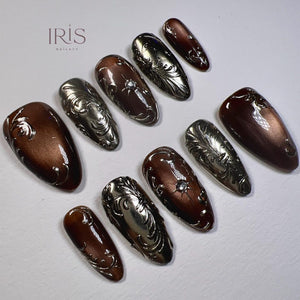A set of press-on nails in various sizes, featuring a metallic silver and brown color scheme. The nails have ornate, embossed designs, including swirls and floral patterns. Some nails have a small, clear gem. The brand name "IRIS NAILART" is visible.