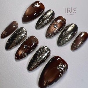 A set of artificial nails in various shapes and sizes. The nails feature a combination of metallic silver and brown hues. Some nails have ornate, raised silver designs, while others are a solid brown color. The brand name "IRIS NAILART" is visible.