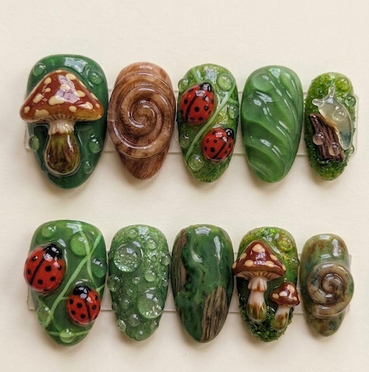 A collection of ten uniquely designed artificial nails. The nails feature various nature-inspired designs, including mushrooms, ladybugs, snails, and leaf patterns. The color palette includes shades of green, brown, and red.