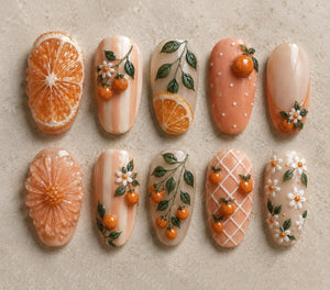 A collection of ten intricately designed nail art samples. The designs feature orange slices, flowers, and miniature oranges, with a color palette of orange, white, and peach. The nails showcase various textures and patterns.