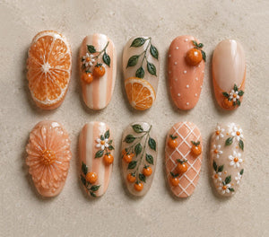 A collection of ten intricately designed nail art pieces. The designs feature orange slices, oranges, flowers, and leaves in shades of orange, white, and green. Some nails have a textured appearance, while others have a smooth, glossy finish.