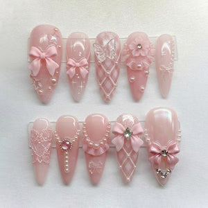 A set of ten artificial nails in a light pink hue, each with unique embellishments. Designs include bows, pearls, lace, and a butterfly, creating a delicate and feminine aesthetic. The nails are displayed on a clear plastic holder.