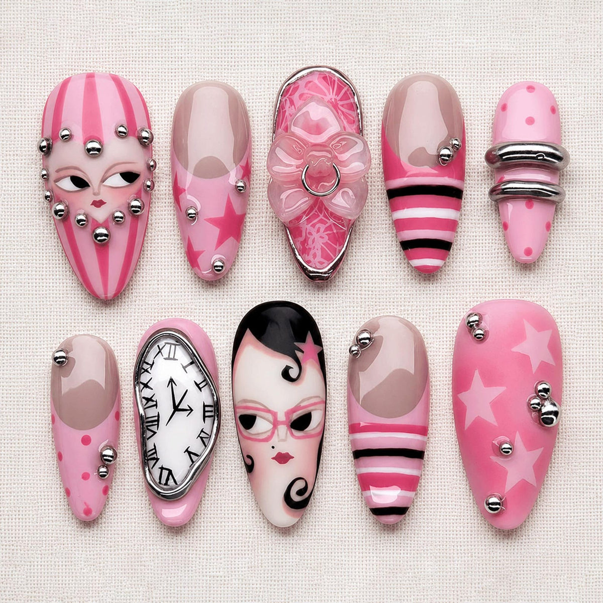 A collection of ten pink and nude-colored artificial nails with various designs. Some nails feature faces, stars, stripes, and a clock face. Embellishments include silver studs and a bow.