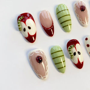 A collection of artificial nails with various designs. Some nails feature a red apple design with a green worm, while others have a pink base with a ladybug. Other nails have green stripes or a pink and red design.