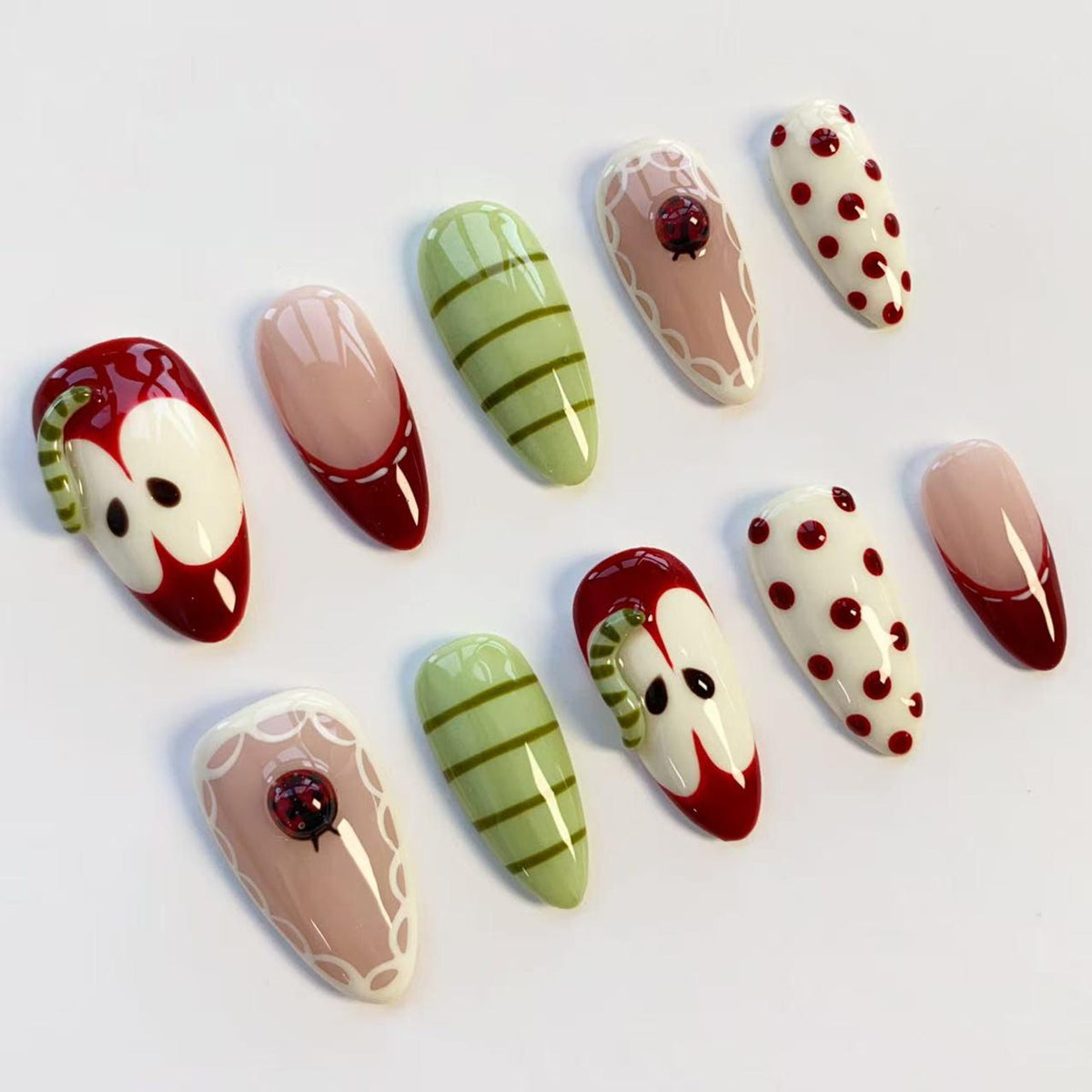 A collection of ten artificial nails with various designs. The nails feature designs such as red apples, green stripes, red polka dots, and a ladybug. Some nails have a nude base with a red tip or white lace.