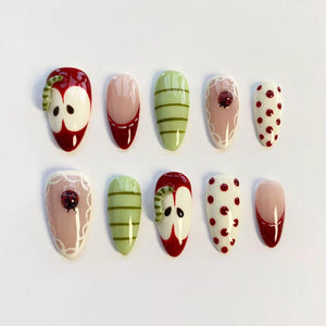 A set of ten artificial nails with various designs. The designs include red apples with worms, pink and red French tips, green striped patterns, ladybugs on pink lace, and white nails with red polka dots. The nails are oval-shaped.