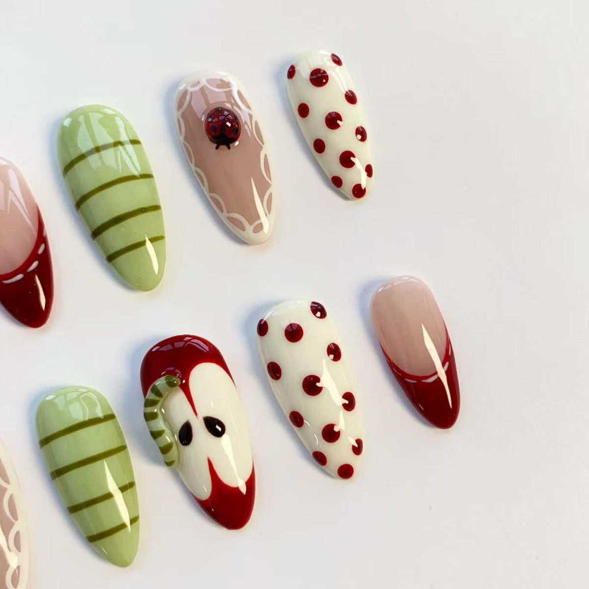 A collection of almond-shaped artificial nails with various designs. Some nails feature red and white polka dots, while others have green stripes. One nail has a ladybug, and another depicts a red apple with a worm.