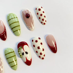 A collection of almond-shaped artificial nails with various designs. Some nails feature red and white polka dots, while others have green stripes. One nail has a ladybug, and another depicts a red apple with a worm.