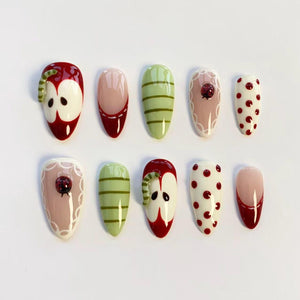 A set of ten almond-shaped false nails with various designs. The designs include red apples with worms, green striped nails, nails with ladybugs, and nails with red polka dots. The nails are painted in a variety of colors including red, green, pink, 
