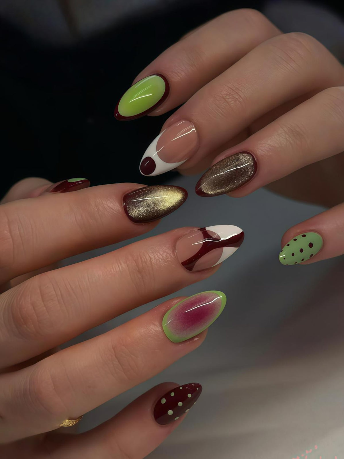 A close-up of a hand with a variety of almond-shaped nail designs. The nails feature colors like green, gold, burgundy, and white, with polka dots and abstract patterns. The designs include a French tip and a gradient effect.