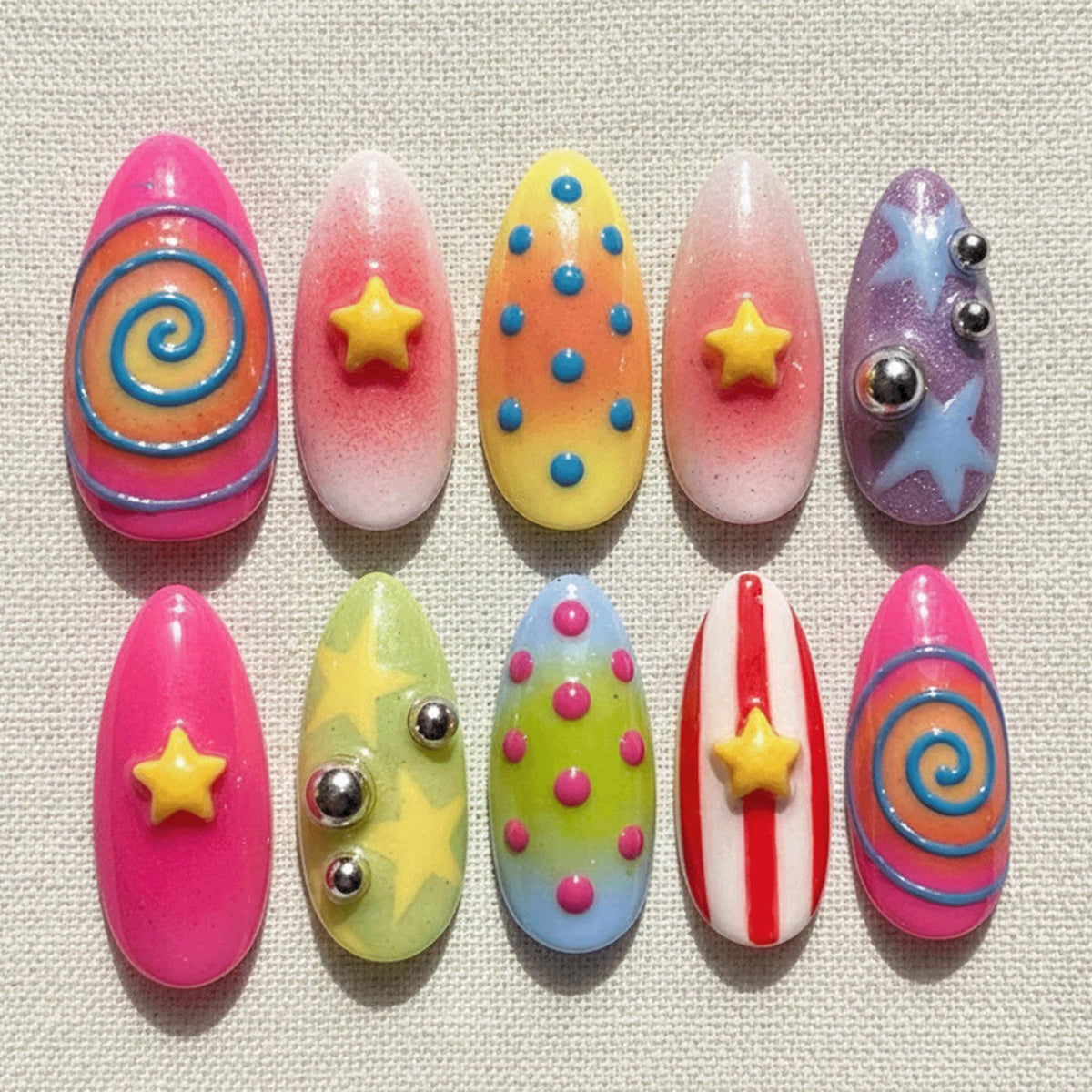 A set of ten artificial nails in various designs. The nails feature bright colors like pink, yellow, and purple, with embellishments such as stars, swirls, dots, and metallic accents. The nails are oval-shaped.