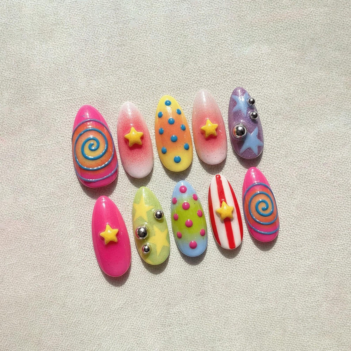 A set of ten artificial nails in various designs. The nails feature bright colors like pink, yellow, and blue, with patterns including swirls, stars, dots, and stripes. Some nails have small silver embellishments.
