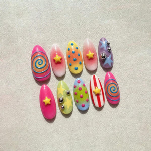 A set of ten artificial nails in various designs. The nails feature bright colors like pink, yellow, and blue, with patterns including swirls, stars, dots, and stripes. Some nails have small silver embellishments.