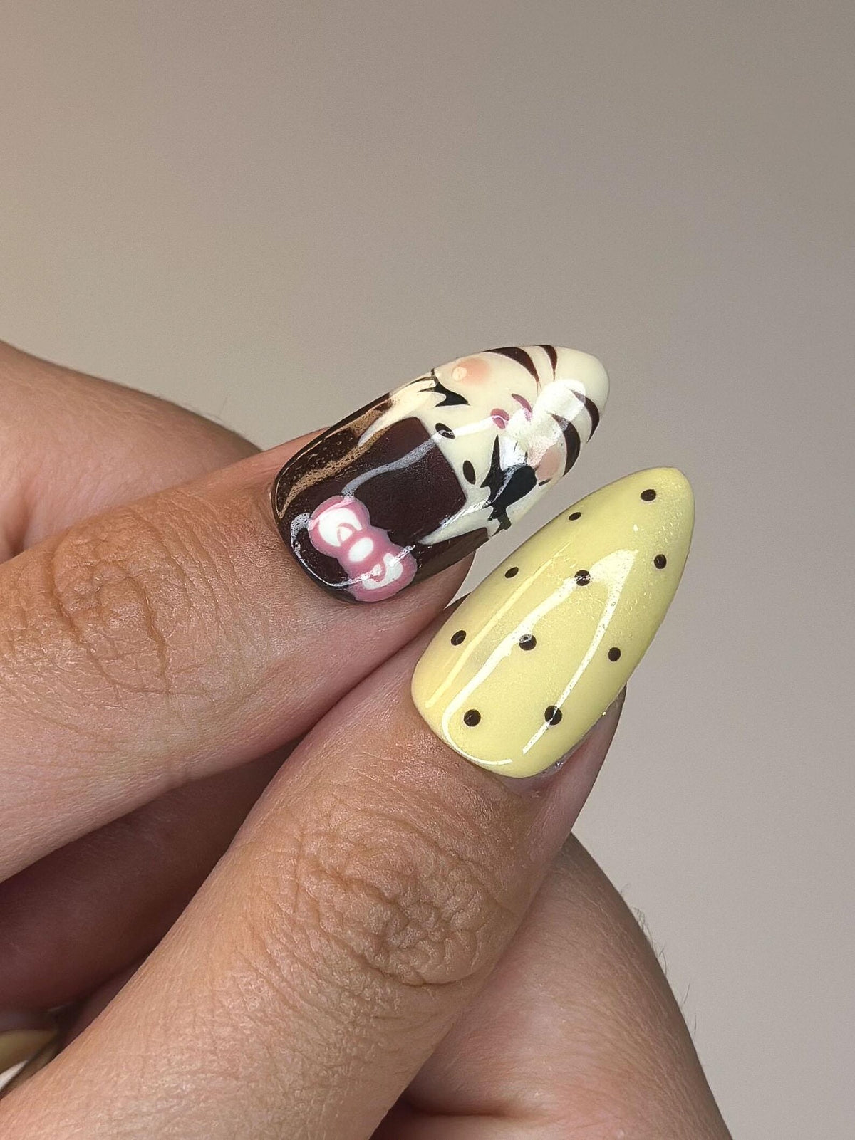 Two manicured nails. One nail features a design with brown, white, and pink elements. The other nail is yellow with small black polka dots. The nails are oval-shaped.