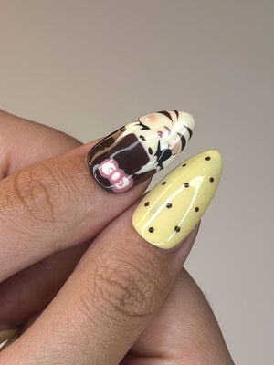 Two manicured nails. One nail features a design with brown, white, and pink elements. The other nail is yellow with small black polka dots. The nails are oval-shaped.