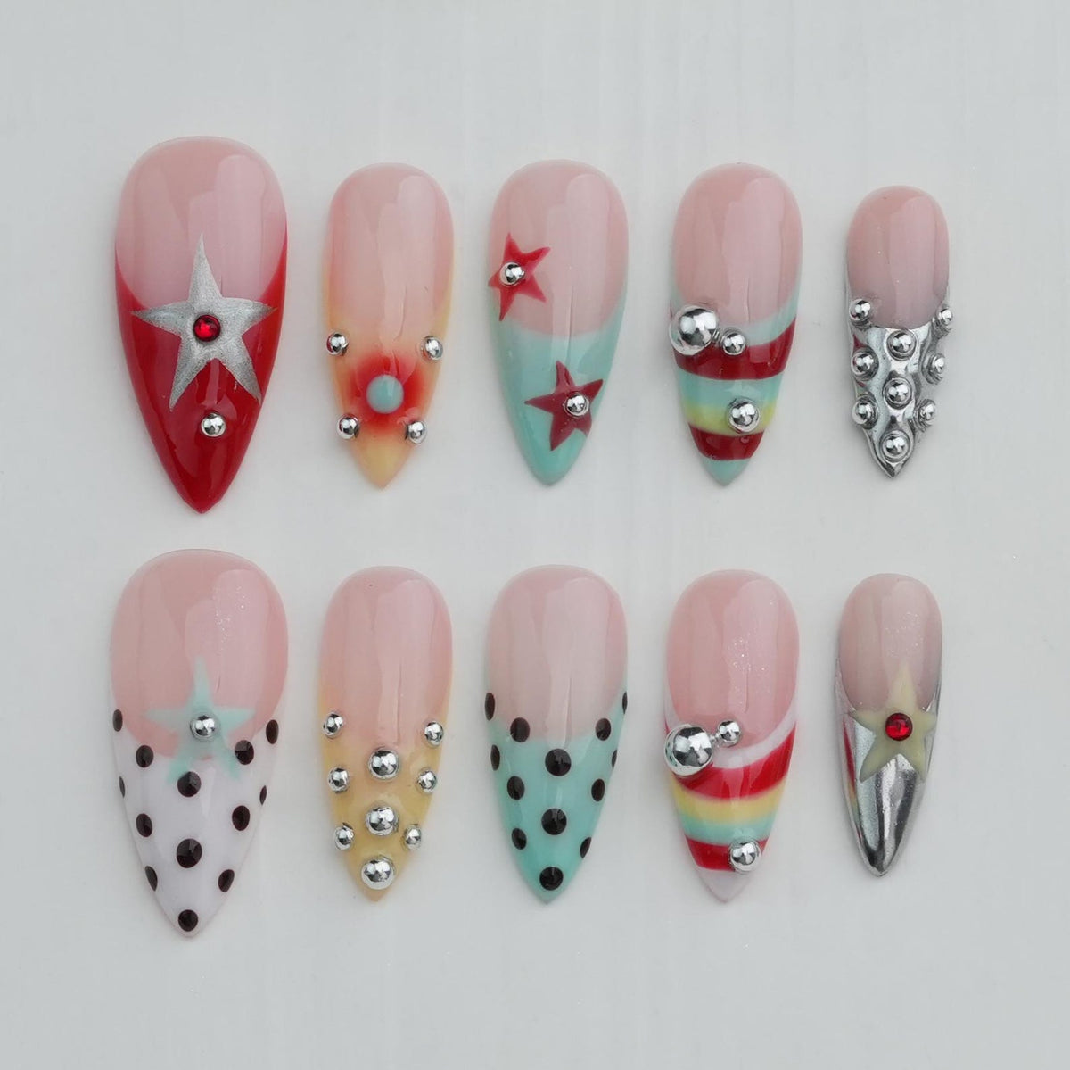 A set of ten almond-shaped artificial nails with various designs. The nails feature a gradient of pink and clear polish, with accents like stars, polka dots, and metallic embellishments in silver and red. Some nails have a red base.