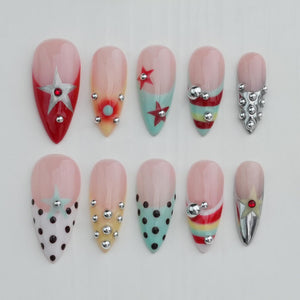 A set of ten almond-shaped artificial nails with various designs. The nails feature a gradient of pink and clear polish, with accents like stars, polka dots, and metallic embellishments in silver and red. Some nails have a red base.