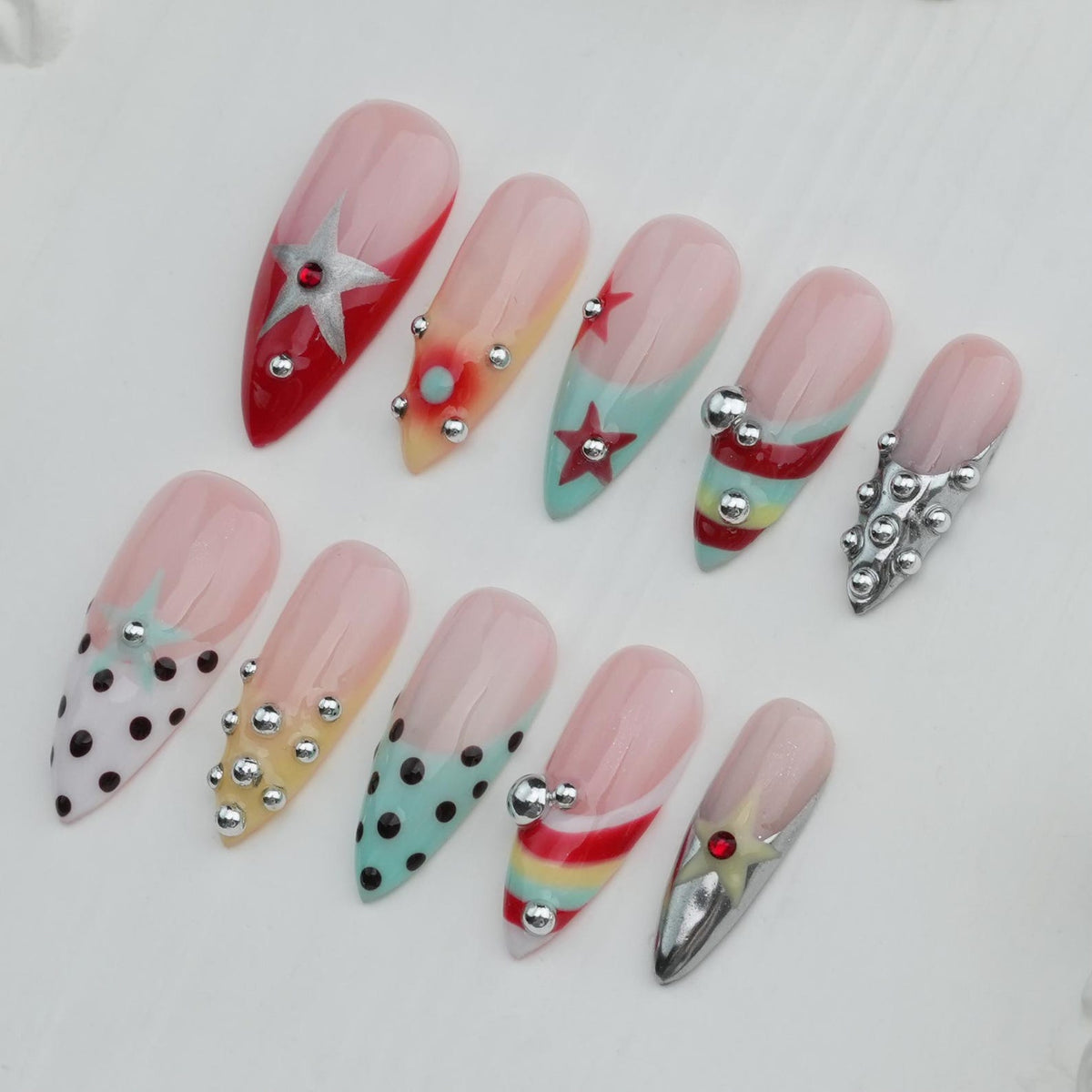 A collection of ten long, almond-shaped artificial nails. Each nail features a different design, including stars, polka dots, and stripes, with accents of silver studs and metallic embellishments. The nails are painted in various colors, including re