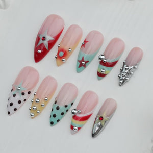 A collection of ten long, almond-shaped artificial nails. Each nail features a different design, including stars, polka dots, and stripes, with accents of silver studs and metallic embellishments. The nails are painted in various colors, including re
