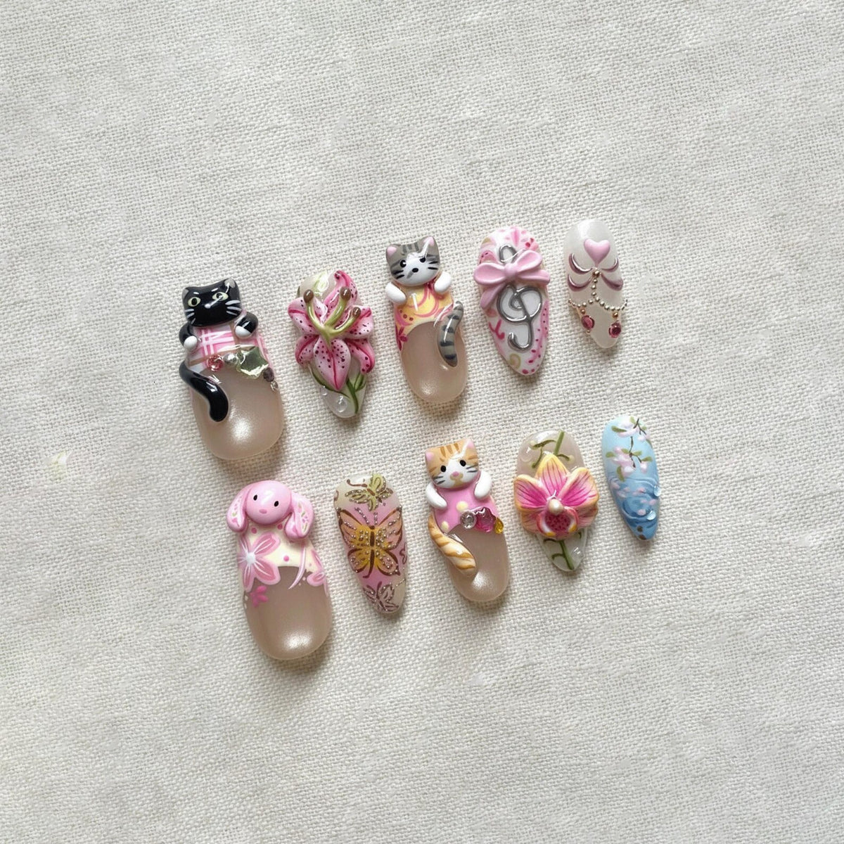 A collection of ten artificial nails, each uniquely designed with 3D embellishments. Designs include a black cat, pink flowers, a gray cat, a pink bow, a heart, a pink dog, a butterfly, an orange cat, pink flowers, and blue flowers.