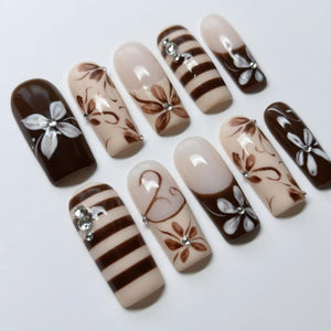 A set of ten artificial nails with various designs. The nails feature a combination of brown, beige, and white colors. Some nails have floral patterns, stripes, or rhinestones. The nails are oval-shaped and have a glossy finish.