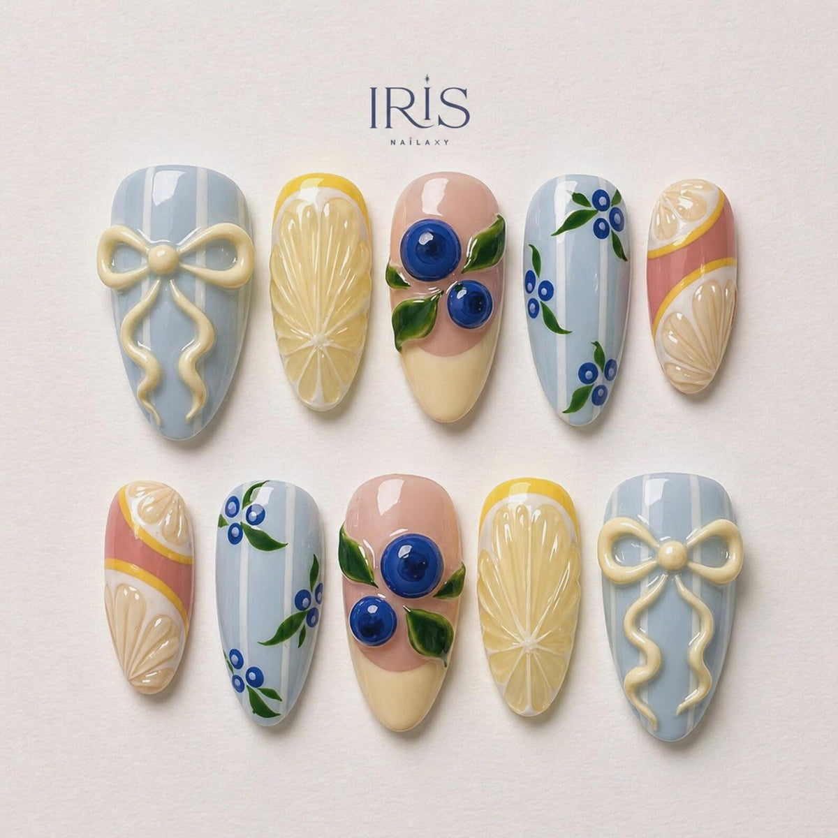 A set of ten almond-shaped artificial nails with various designs. The designs include light blue stripes with a bow, lemon slices, blueberries with green leaves, and pink and yellow patterns. The brand name "IRIS NAILARY" is at the top.