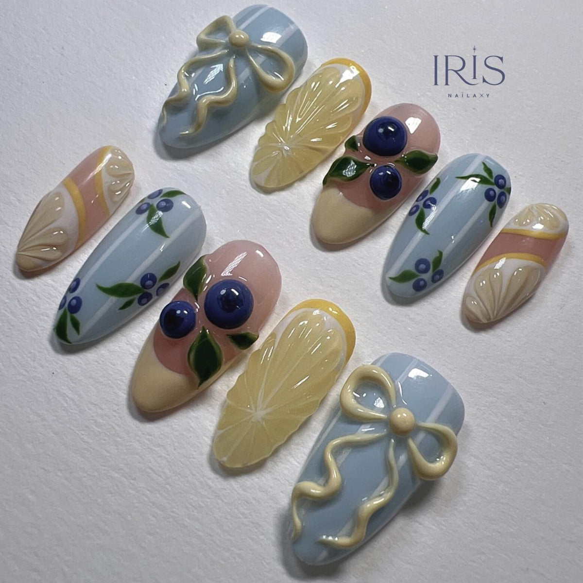 A set of ten almond-shaped artificial nails with various designs. The designs include blue bows, lemon slices, blueberries with green leaves, and blue and white striped patterns. The nails are painted in pastel colors with the text "IRIS NAILART".