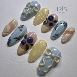 A set of ten almond-shaped artificial nails with various designs. The designs include blue bows, lemon slices, blueberries with green leaves, and blue and white striped patterns. The nails are painted in pastel colors with the text "IRIS NAILART".