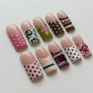 A collection of ten artificial nails with various designs. The nails feature a natural pink tip and are decorated with patterns like polka dots, floral designs, and plaid. Some nails have rhinestones for added detail. The color palette includes pink,