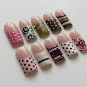 A collection of ten artificial nails with various designs. The nails feature a natural pink tip and are decorated with patterns like polka dots, floral designs, and stripes. Some nails have rhinestones for added detail. The nails are set against a wh