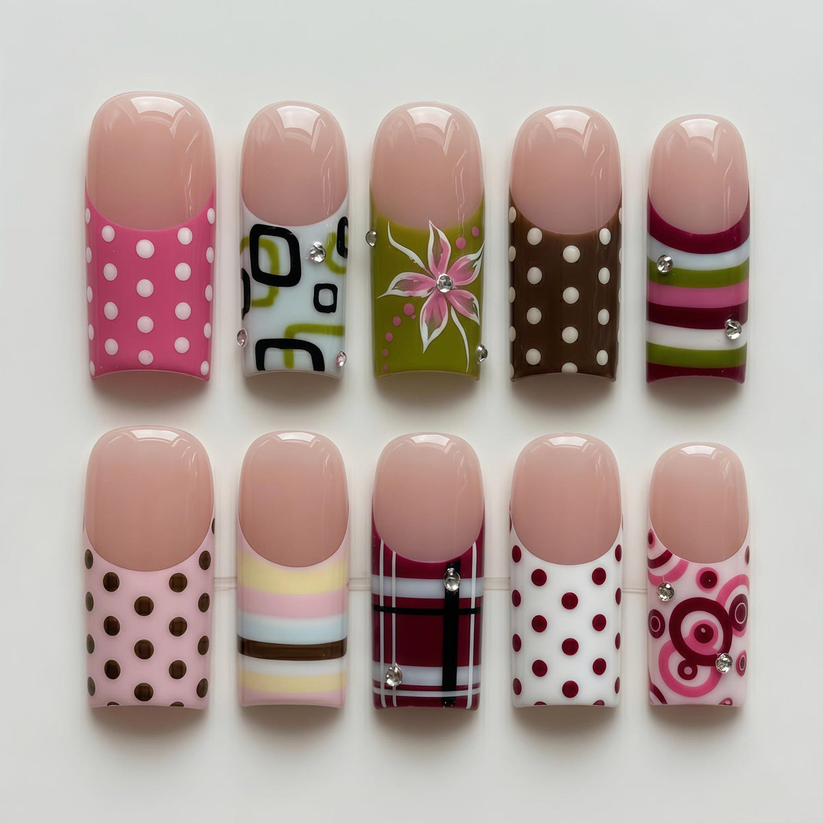 A set of ten artificial nails with various designs. The nails feature a natural pink tip and a range of patterns, including polka dots, geometric shapes, floral designs, and stripes. Some nails have decorative rhinestones.
