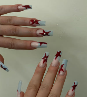 Long, square-shaped acrylic nails with a light blue and nude gradient. Each nail features a dark red star design and a small silver stud. The nails are well-manicured and showcase a modern nail art style.