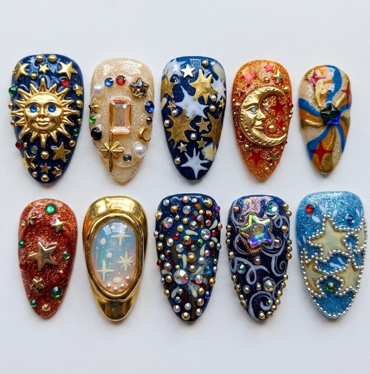 A collection of ten intricately designed artificial nails. Each nail features unique celestial-themed embellishments, including suns, moons, stars, and various gemstones. The color palette includes blues, golds, reds, and iridescent accents, creating