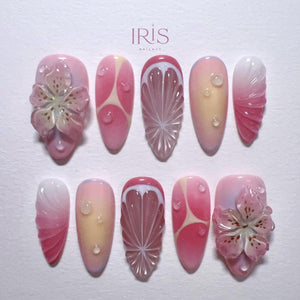 A set of ten artificial nails with various designs. The nails feature a gradient of pink and white, with some adorned with 3D flower and fruit slice designs. Some nails have water droplet accents. The brand name "IRIS" is at the top.