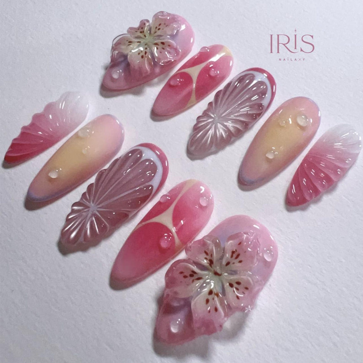 A collection of ten long, almond-shaped artificial nails. The nails feature a variety of designs, including floral embellishments, fruit slice patterns, and gradient color schemes in shades of pink, white, and yellow. The text "IRIS NAIL ART" is visi
