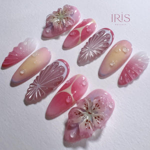 A collection of ten long, almond-shaped artificial nails. The nails feature a variety of designs, including floral embellishments, fruit slice patterns, and gradient color schemes in shades of pink, white, and yellow. The text "IRIS NAIL ART" is visi